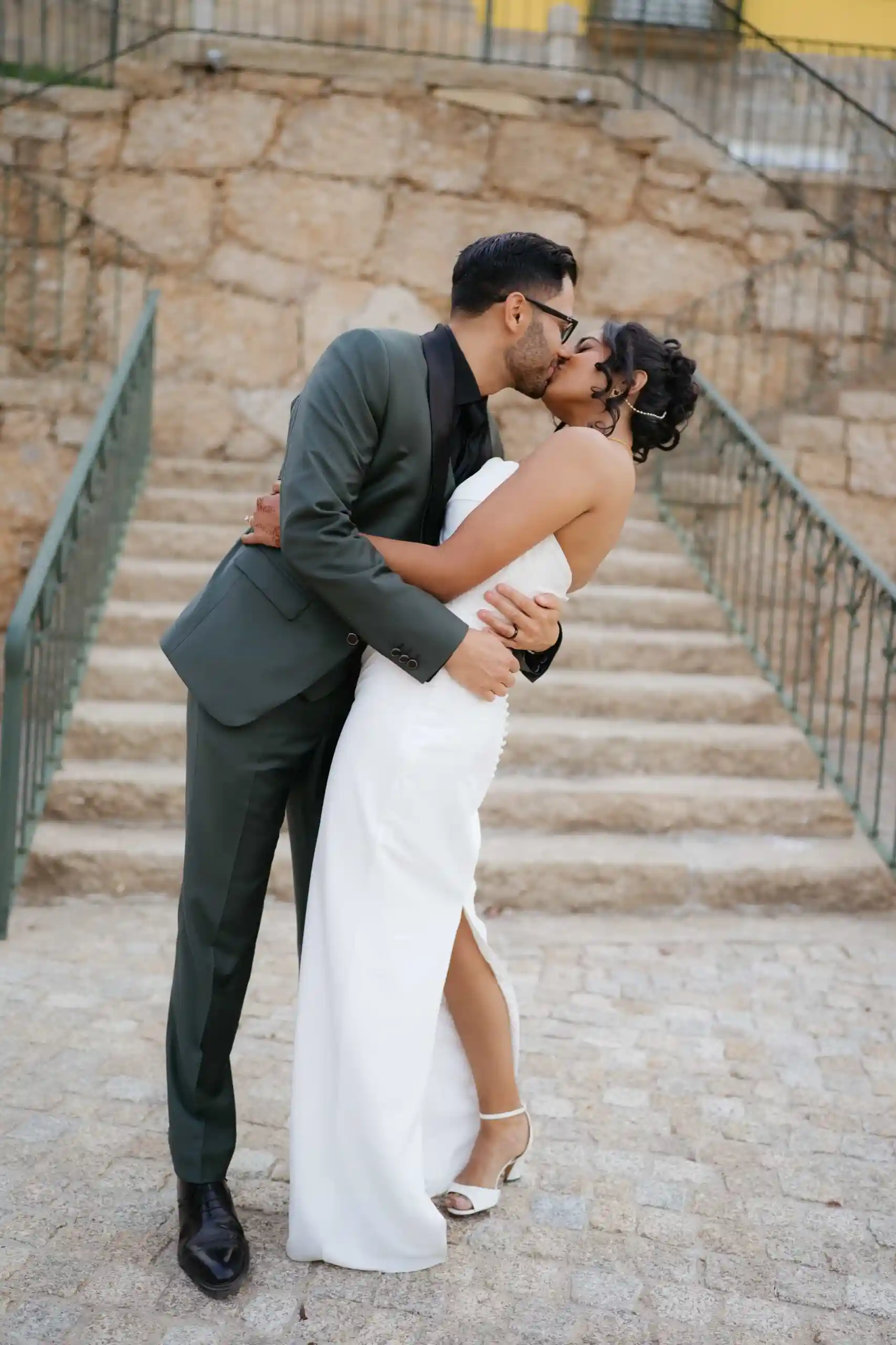 Groom in a dark green suit kissing a bride in a white wedding dress on stone stairs at an outdoor wedding venue in Portugal.